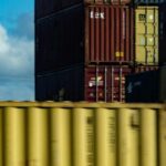 A stack of colorful shipping containers at a port, symbolizing international trade and business regulations, with a blurred yellow container in the foreground. The image represents the impact of the Foreign Business Act in Thailand on foreign trade and investment.