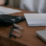 Person working on accounting in Thailand at a wooden desk with a calculator, notebook, and laptop.