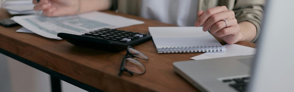 Person working on accounting in Thailand at a wooden desk with a calculator, notebook, and laptop.