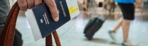 A close-up of a person holding a U.S. passport and boarding pass at an airport he has all the DTV Visa Requirements, with blurred travelers in the background.