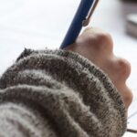 A person in a sweater writing on a document with a pen, symbolizing education, visa applications, or academic paperwork.