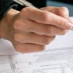 A person filling out a corporate income tax in Thailand form with a pen, focusing on financial documents on a wooden desk.