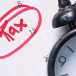 Календарь с выделенной красной ручкой датой "14" и пометкой "Tax", а также черный будильник, подчеркивающий срочность уплаты налоги в Таиланде.