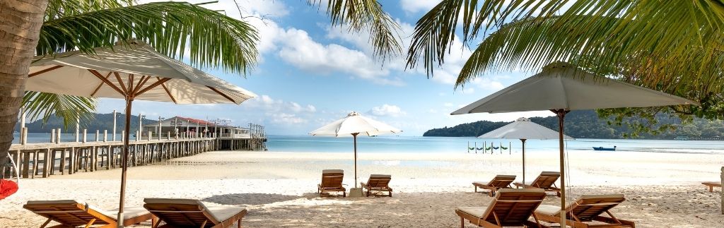 Sonnenliegen und Sonnenschirme am tropischen Strand mit Palmen und Steg – idealer Ort für Auswanderer in Thailand.