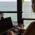 Woman working remotely from a beach in Thailand using laptop under wooden terrace, illustrating Thailand remote work visa lifestyle for digital nomads