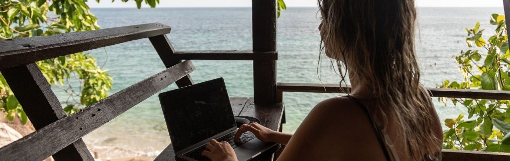 Woman working remotely from a beach in Thailand using laptop under wooden terrace, illustrating Thailand remote work visa lifestyle for digital nomads